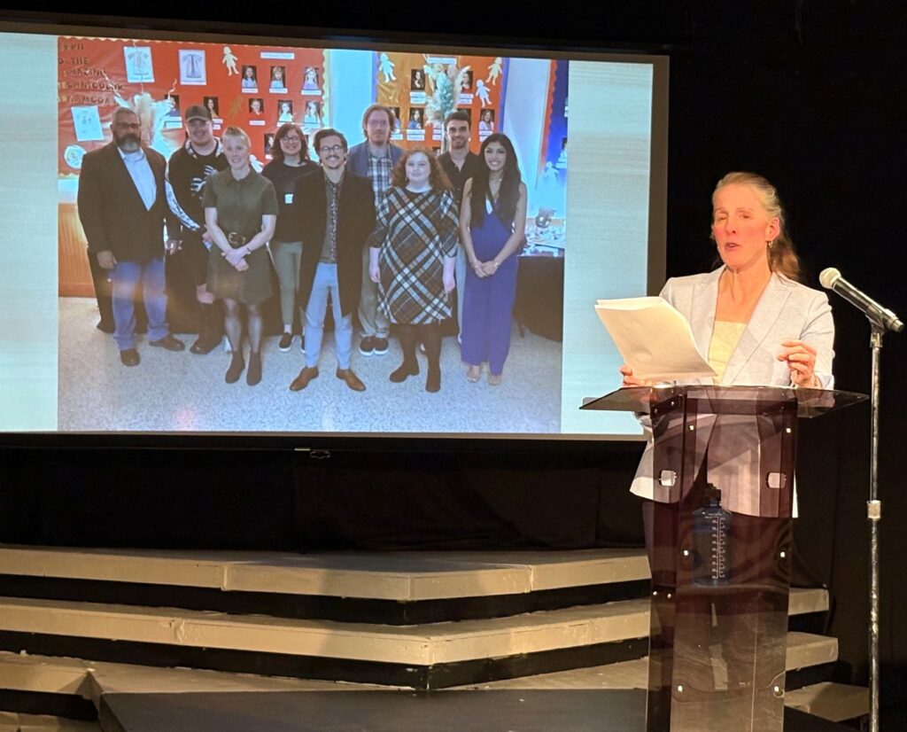 Kelsey Trom speaks as a photo with Tusculum students and faculty displays in the background.