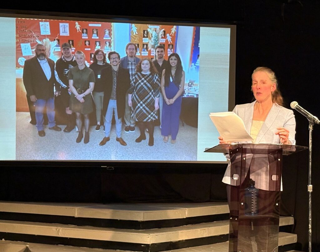 Kelsey Trom speaks as a photo with Tusculum students and faculty displays in the background.