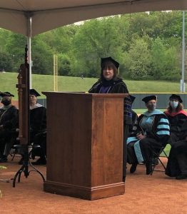 Dr. Hunsader wearing graduation attire, speaking at a podium 
