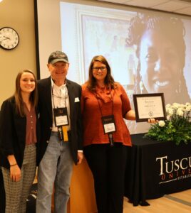 Left to right, Belle Kemp, Bob Kleinertz and Tiffany Greer honor Erika Witt, who participated virtually in the ceremony and can be seen on the screen.